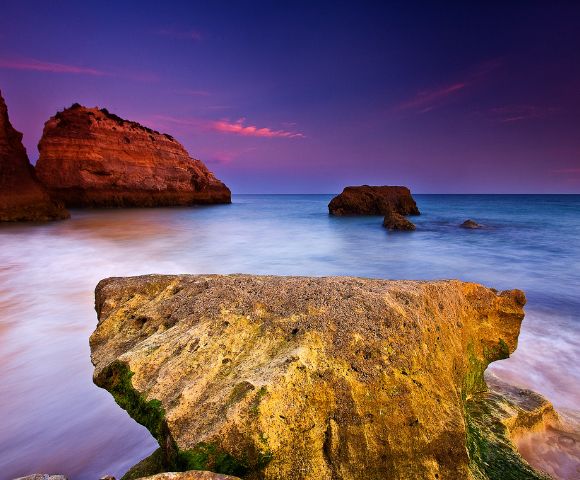A large, textured rock dominates the foreground against a blurred seascape at dusk, with vibrant purple and orange skies and distant cliffs enhancing tranquility.