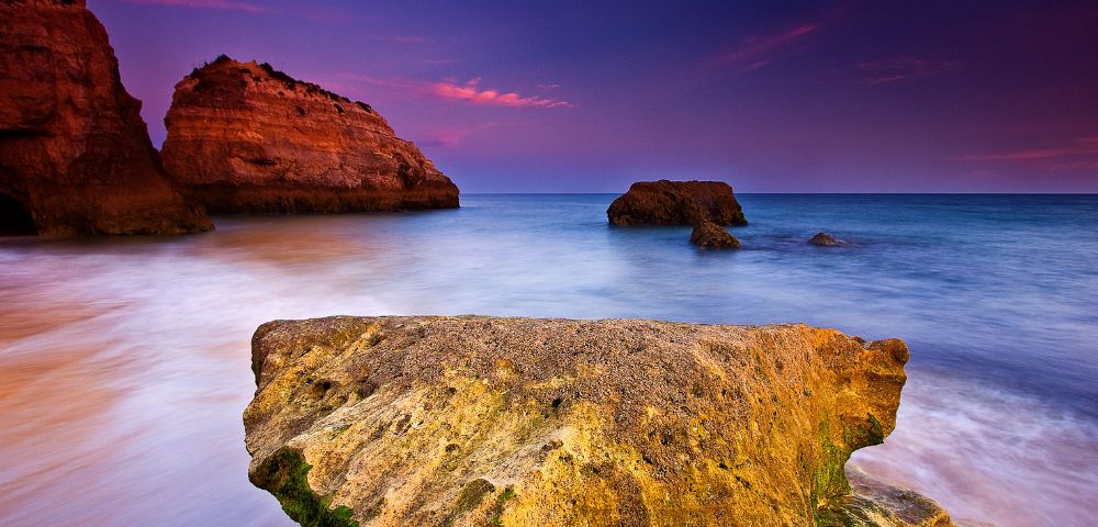 A large, textured rock dominates the foreground against a blurred seascape at dusk, with vibrant purple and orange skies and distant cliffs enhancing tranquility.