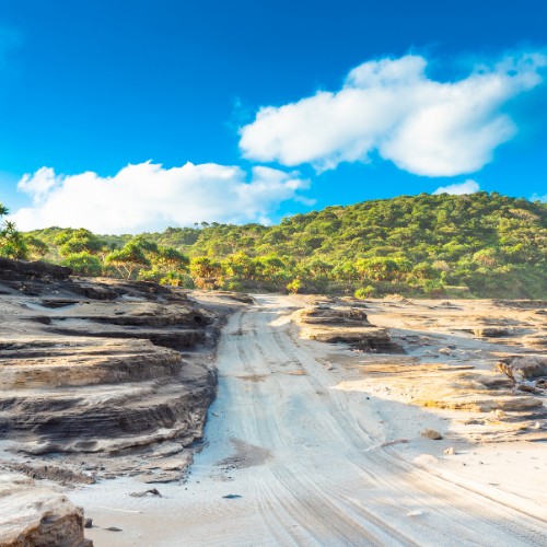 Sandy path with rugged rock formations leads to lush green hills under a bright blue sky with fluffy clouds.