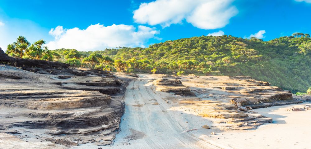 Sandy path with rugged rock formations leads to lush green hills under a bright blue sky with fluffy clouds.