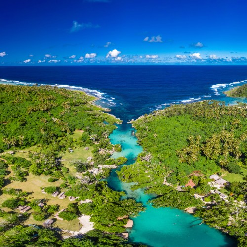 Aerial view of a tropical lagoon with vivid turquoise water flowing into the ocean, surrounded by lush green forest, under a bright blue sky.