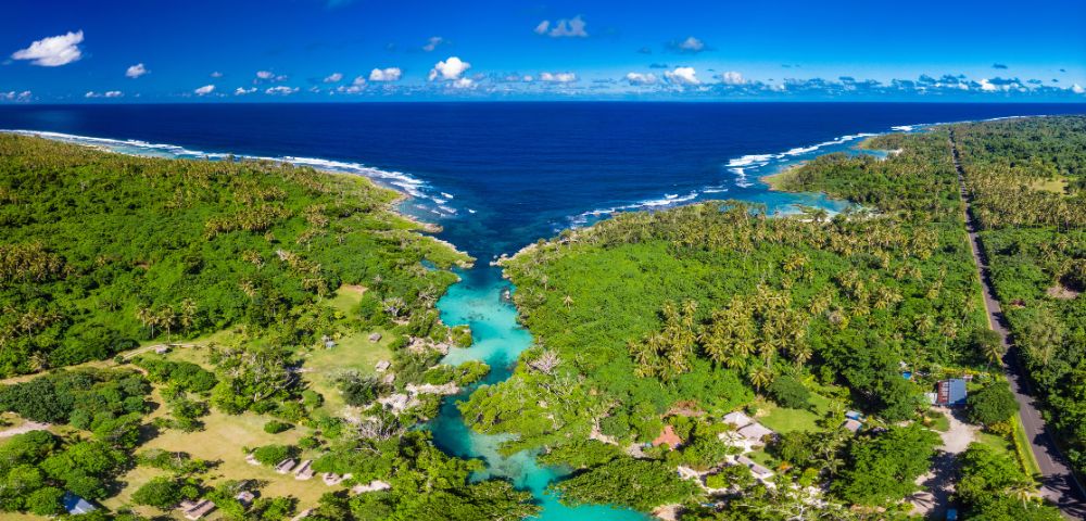 Aerial view of a tropical lagoon with vivid turquoise water flowing into the ocean, surrounded by lush green forest, under a bright blue sky.