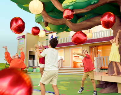 Children play joyfully under a colorful tree adorned with shiny red balloons, near a vibrant, welcoming house.