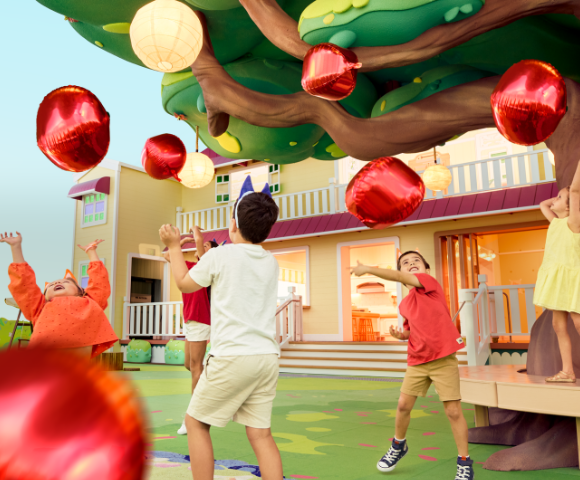 Children play joyfully under a colorful tree adorned with shiny red balloons, near a vibrant, welcoming house.