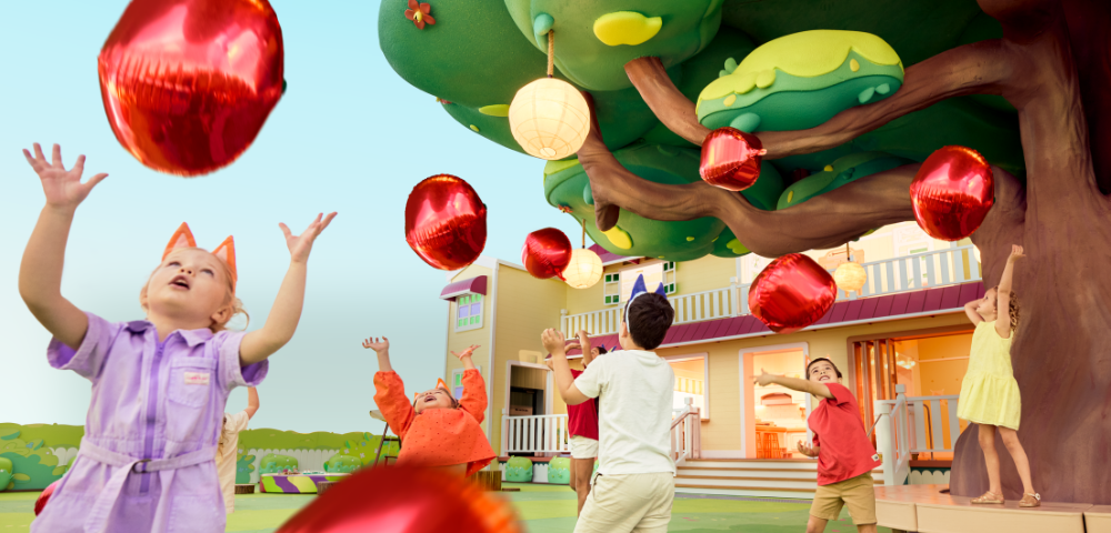Children play joyfully under a colorful tree adorned with shiny red balloons, near a vibrant, welcoming house.
