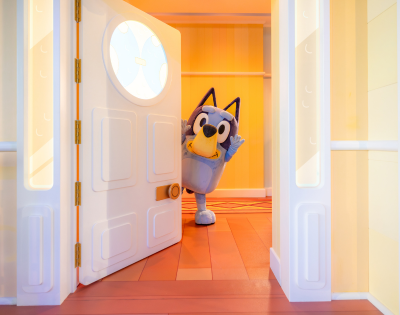 Bluey, a cartoon dog, peeks playfully from behind a partially open door in a colorful room.
