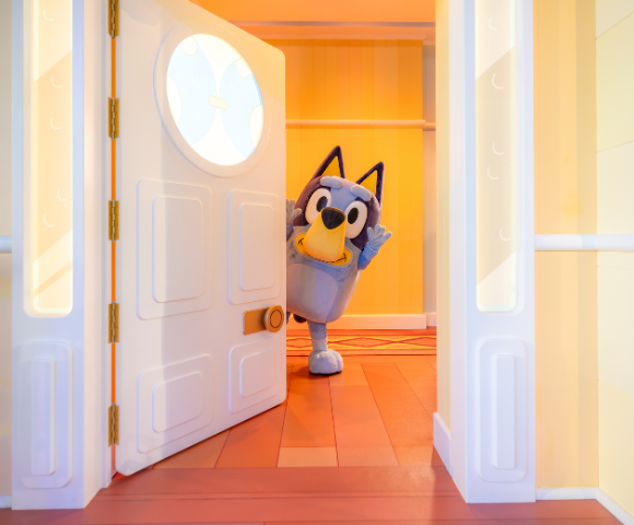 Bluey, a cartoon dog, peeks playfully from behind a partially open door in a colorful room.