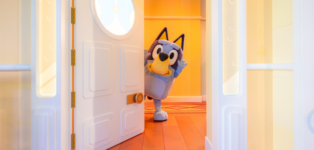 Bluey, a cartoon dog, peeks playfully from behind a partially open door in a colorful room.
