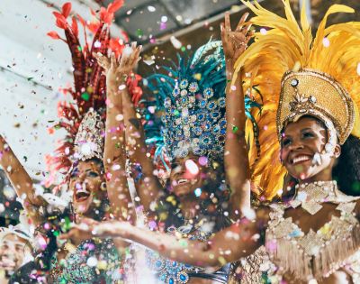 Three dancers in vibrant feathered costumes celebrate joyfully, surrounded by colorful confetti. Radiant smiles create an energetic, festive atmosphere.
