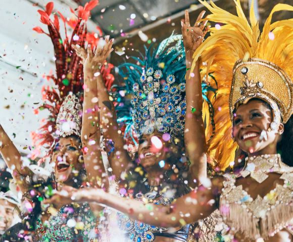 Three dancers in vibrant feathered costumes celebrate joyfully, surrounded by colorful confetti. Radiant smiles create an energetic, festive atmosphere.
