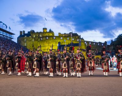 A large group dressed in traditional Scottish attire poses in front of Edinburgh Castle illuminated in green.