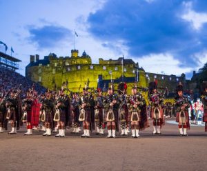A large group dressed in traditional Scottish attire poses in front of Edinburgh Castle illuminated in green.