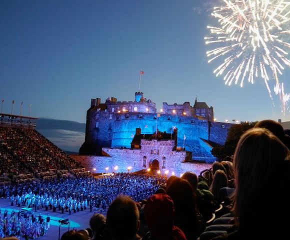Edinburgh Castle glows at night with fireworks overhead and a crowd gathered below.