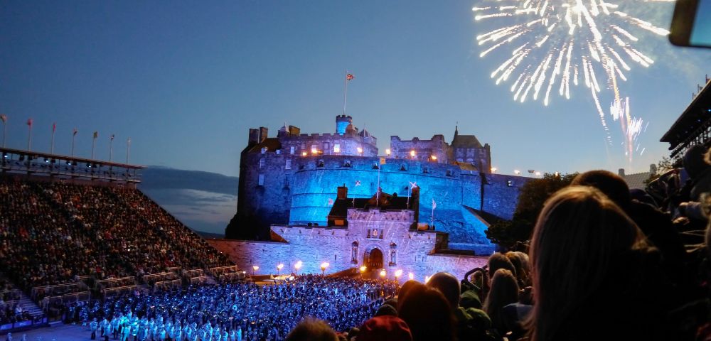 Edinburgh Castle glows at night with fireworks overhead and a crowd gathered below.