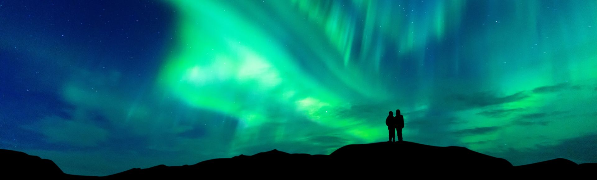 A couple stands on a hill, silhouetted against the vibrant aurora borealis lighting up the night sky.
