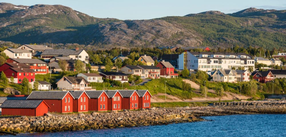 Scenic coastal view with red wooden houses beside the water, framed by green hills and distant mountains under a clear sky.