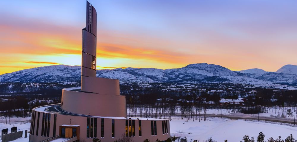 Modern church building with a spiral design against a colorful sunset and snow-covered mountains in the background.