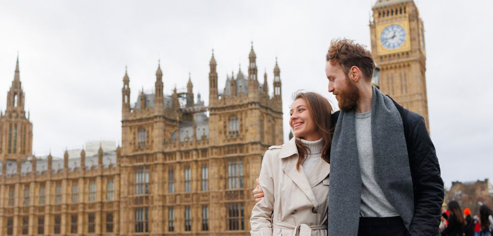 A couple poses together on a bridge in front of the iconic Houses of Parliament and Big Ben in London, under a cloudy sky.