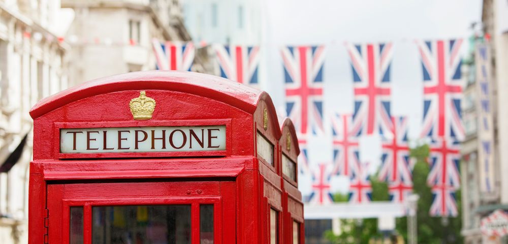 A classic red British telephone booth stands in the foreground, decorated with Union Jack flags overhead in a city street.