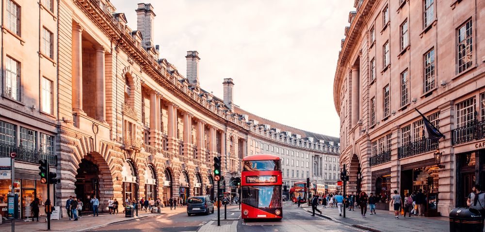 A busy London street with a red double-decker bus, historic buildings, and pedestrians under a warm sunset glow.