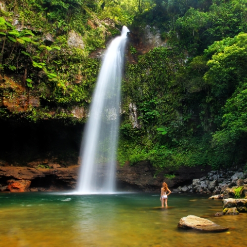 A woman stands in a serene pool at the base of a tall, cascading waterfall surrounded by lush green foliage, evoking a sense of tranquility and awe.