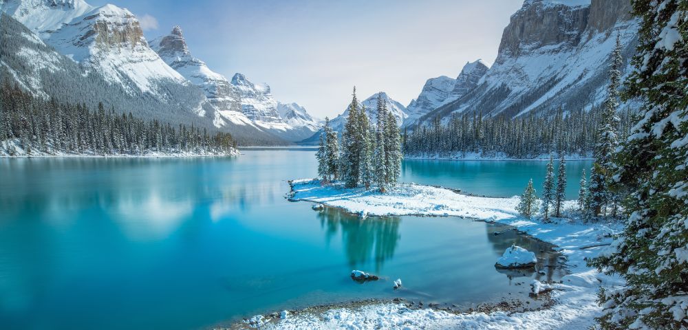 A serene winter landscape featuring a turquoise lake, snow-covered trees, and majestic mountains under a clear sky in Jasper National Park, Canada.