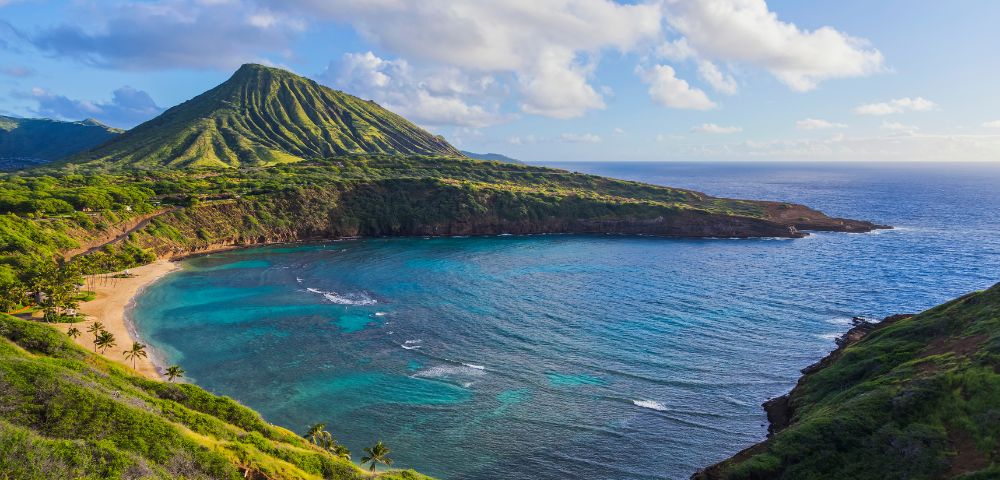 Serene coastal landscape of a turquoise bay with lush green mountains in the background under a partly cloudy sky, conveying tranquility and natural beauty.
