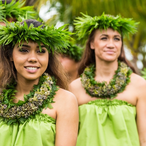 Two women in green dresses and leaf crowns stand outdoors, smiling. They appear relaxed and joyful, with palm trees in the blurred background.