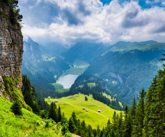 A breathtaking mountainous landscape with a lush green valley, steep rocky cliff on the left, dense evergreen trees on the right, rolling green hills with small houses, a winding path leading to a serene lake, and towering mountains partially covered by clouds in the background.