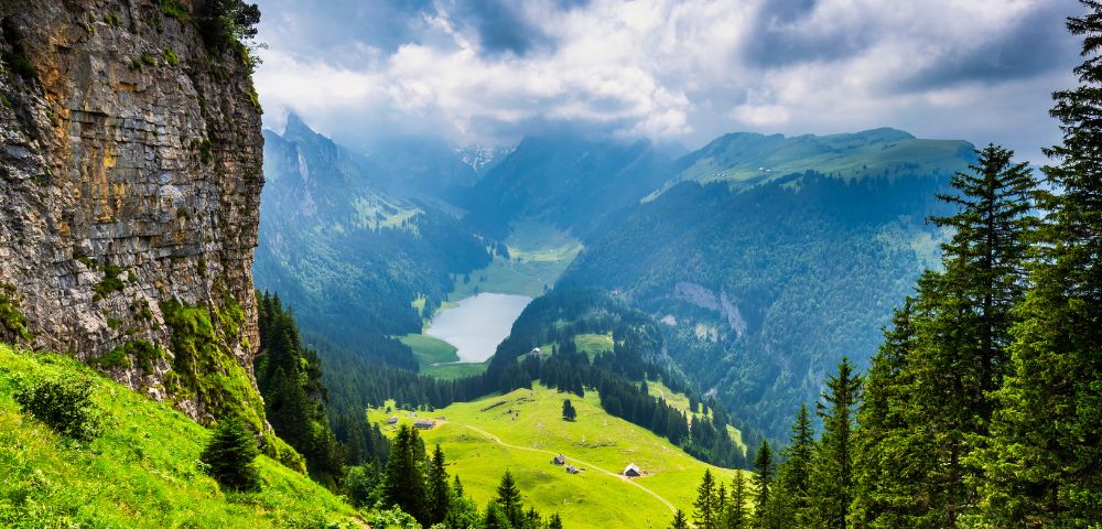 A breathtaking mountainous landscape with a lush green valley, steep rocky cliff on the left, dense evergreen trees on the right, rolling green hills with small houses, a winding path leading to a serene lake, and towering mountains partially covered by clouds in the background.