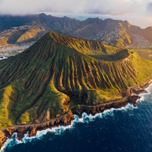 Aerial view of a lush, green volcanic mountain with deep ridges surrounded by ocean waves and a winding coastal road, conveying a serene, majestic atmosphere.