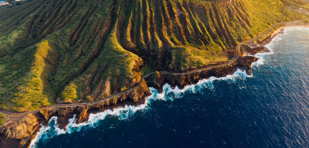 Aerial view of a lush, green volcanic mountain with deep ridges surrounded by ocean waves and a winding coastal road, conveying a serene, majestic atmosphere.