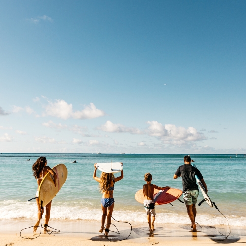 Four people holding surfboards walk towards the ocean on a sunny beach day. The sky is clear with few clouds, creating a joyful and adventurous vibe.