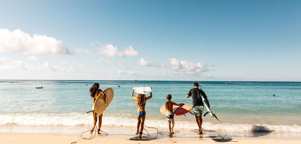 Four people holding surfboards walk towards the ocean on a sunny beach day. The sky is clear with few clouds, creating a joyful and adventurous vibe.