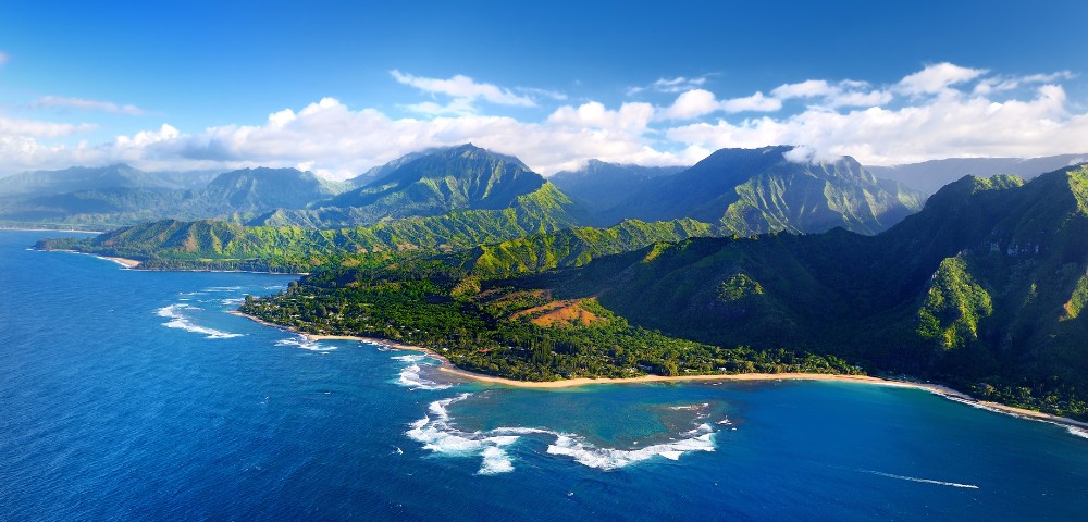 Aerial view of a scenic coastal landscape with lush green mountains, clear blue ocean waters, white waves, and a partly cloudy sky, conveying tranquility.