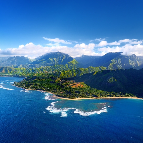 Aerial view of a scenic coastal landscape with lush green mountains, clear blue ocean waters, white waves, and a partly cloudy sky, conveying tranquility.