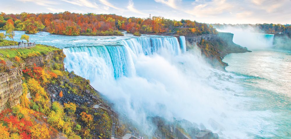 Niagara Falls cascades powerfully over cliffs, surrounded by vibrant autumn foliage and a misty river under a blue sky.