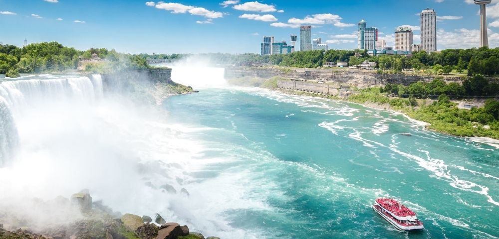 A scenic view of Niagara Falls with a sightseeing boat on the river, framed by lush green trees and a vibrant city skyline under a blue sky.