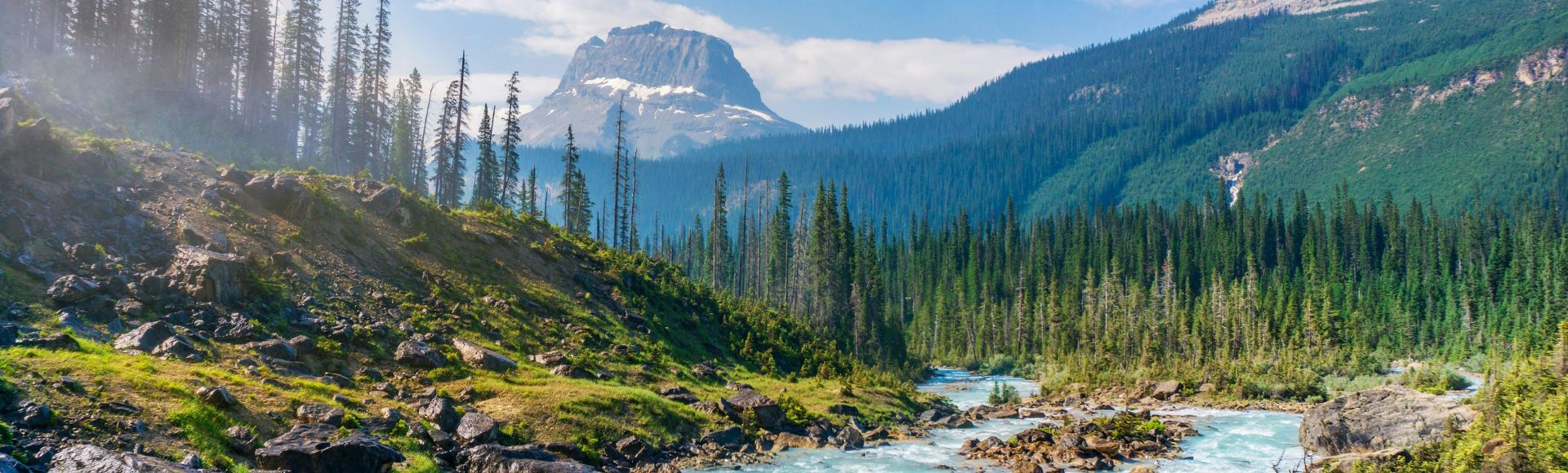 A picturesque landscape featuring a flowing turquoise river, rocky banks, and alpine forest with a mountain in the background under a blue sky.