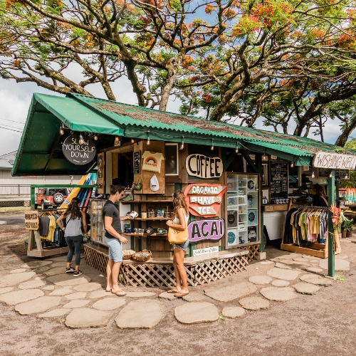 Small, vibrant outdoor shop with a green roof, displaying signs for coffee, organic shave ice, and acai. People browse souvenirs under lush trees, conveying a tropical, casual vibe.