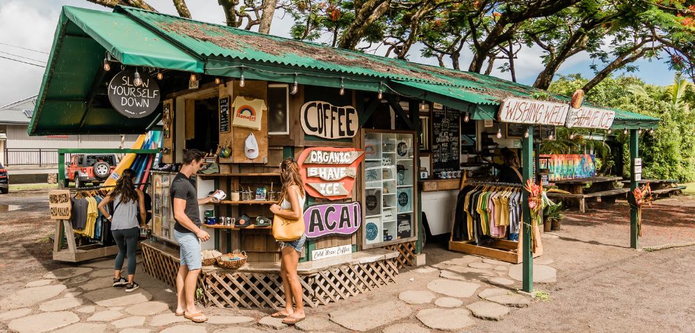 Small, vibrant outdoor shop with a green roof, displaying signs for coffee, organic shave ice, and acai. People browse souvenirs under lush trees, conveying a tropical, casual vibe.