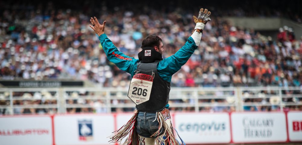 A rodeo cowboy in bright clothing raises his arms in triumph, surrounded by a cheering crowd and sandy arena at the Calgary Stampede.