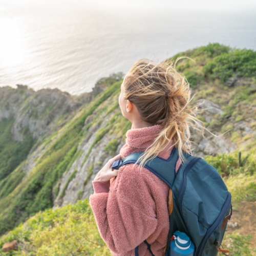 A woman with a backpack stands on a cliffside, overlooking the ocean. Her hair is windswept, and she wears a pink jacket, conveying a sense of adventure.
