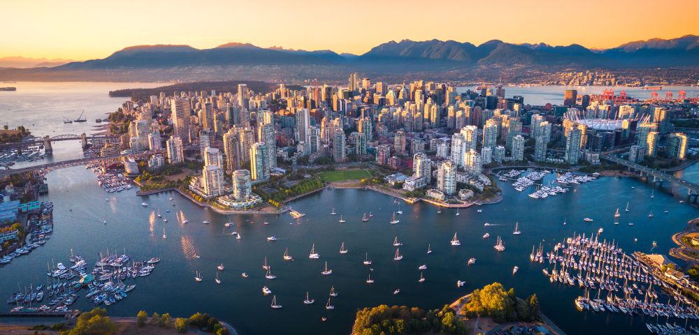 A vibrant aerial view of Vancouver at sunset, showcasing skyscrapers, boats in the harbor, and the backdrop of mountains.