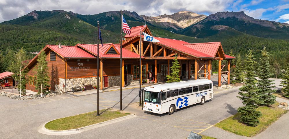 A wooden lodge named Denali Princess Wilderness Lodge with a red roof and a bus parked outside, surrounded by lush mountains and trees.