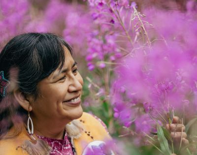 A woman stands among blooming pink flowers, wearing colorful traditional attire and gazing thoughtfully at her surroundings.