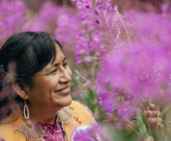 A woman stands among blooming pink flowers, wearing colorful traditional attire and gazing thoughtfully at her surroundings.