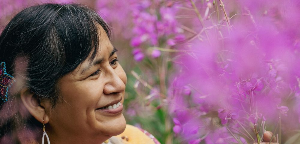 A woman stands among blooming pink flowers, wearing colorful traditional attire and gazing thoughtfully at her surroundings.