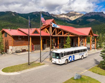 A wooden lodge named Denali Princess Wilderness Lodge with a red roof and a bus parked outside, surrounded by lush mountains and trees.