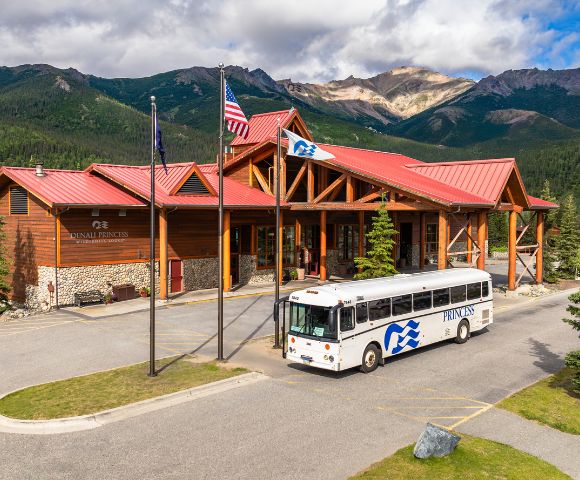 A wooden lodge named Denali Princess Wilderness Lodge with a red roof and a bus parked outside, surrounded by lush mountains and trees.